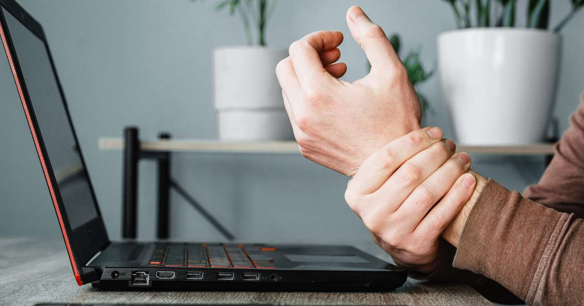 Un homme avec un pull brun se tient le poignet. On devine une douleur.